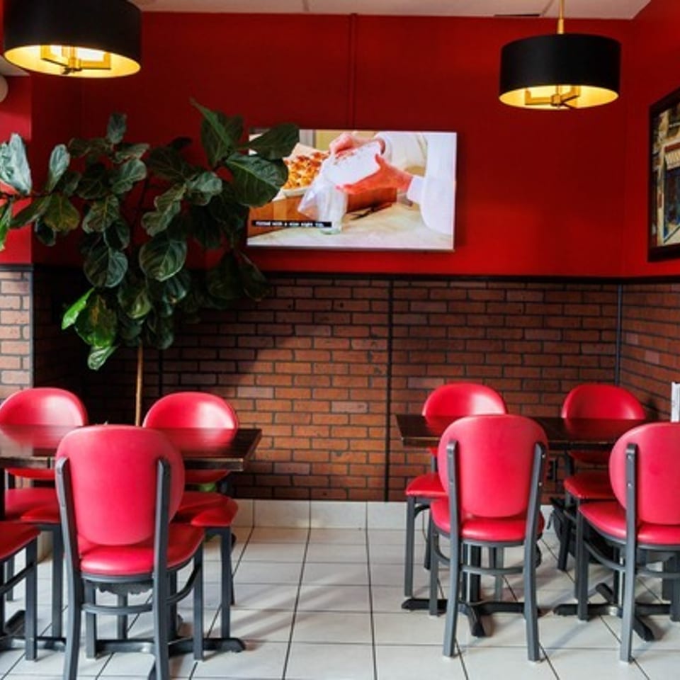Restaurant interior with red walls, brick wainscoting, red chairs, dark tables, and a TV showing food.