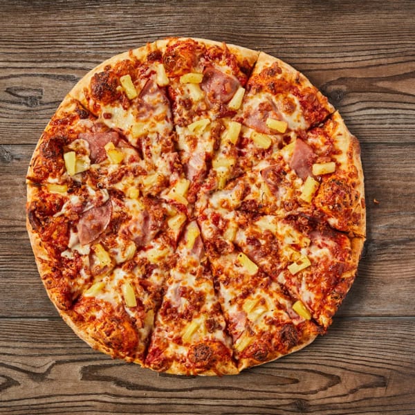 A whole Hawaiian pizza, cut into slices, with ham and pineapple on a wooden table.