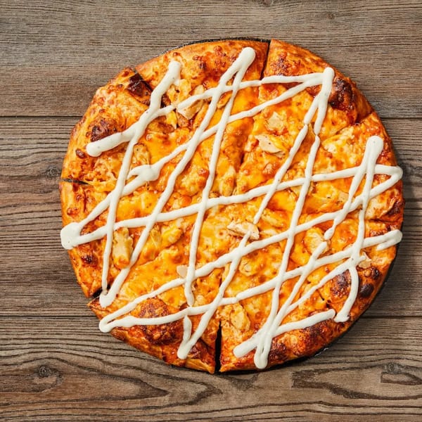 Overhead view of a sliced chicken pizza with white sauce drizzled on a wooden table.