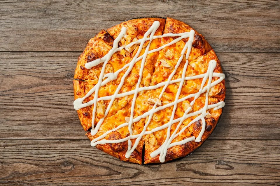 Overhead view of a sliced chicken pizza with white sauce drizzled on a wooden table.