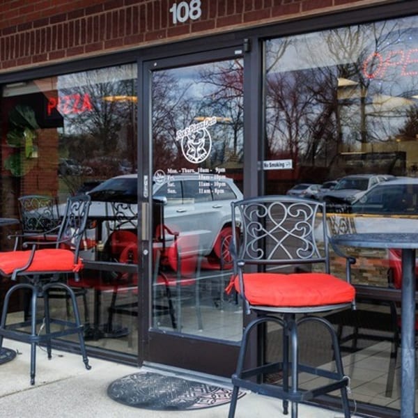Outdoor patio seating with black metal tables and chairs with red cushions in front of a brick building.